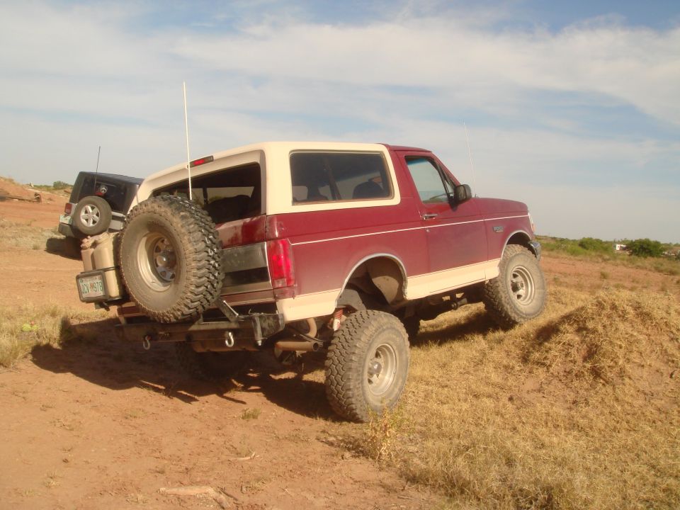Red and tan Ford Bronco with spare tire and jerry can on a dirt hill, black Jeep in background.