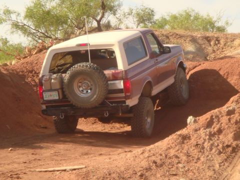 1992 Ford Bronco - A maroon and cream 1992 Ford Bronco is driving off-road on a dirt path, with a spare tire mounted on the back.
