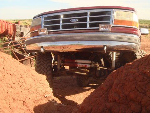 1992 Ford Bronco - The front end of a lifted, maroon Ford Bronco is shown angled down into a dirt ditch, with its chrome bumper and grille prominently display…