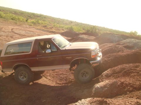 1992 Ford Bronco - A maroon and tan Ford Bronco is shown from the side, driving on a dirt trail with hills in the background. The vehicle is angled upwards, w…