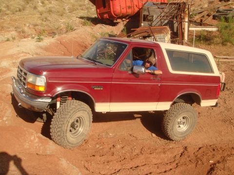 1992 Ford Bronco - A red 1992 Ford Bronco with a tan top is parked on a dirt path with a person leaning out the driver's window. The vehicle has large off-roa…
