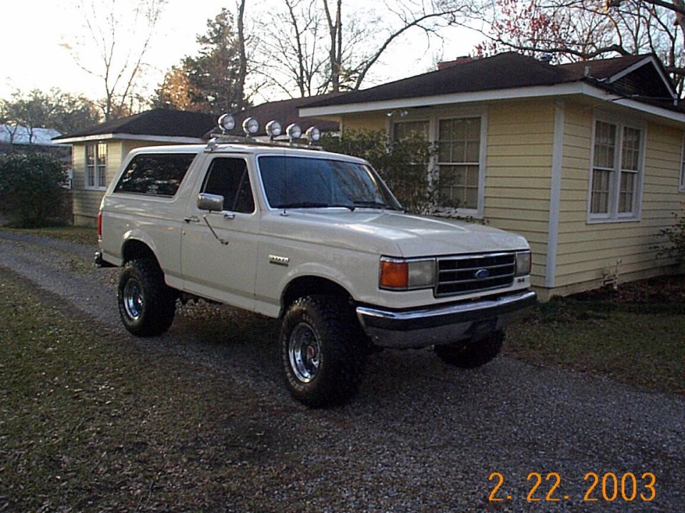 White 1990 Ford Bronco parked on gravel driveway in front of a yellow house, has roof-mounted lights