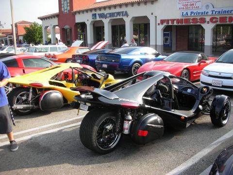 1998 Ford Crown Victoria - black Campagna T-Rex three-wheeled vehicle parked among sports cars and a yellow motorcycle at a car meet.