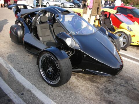 1998 Ford Crown Victoria - black three-wheeled tandem roadster with a single rear wheel and roll bar sits in a parking lot beside sports cars.