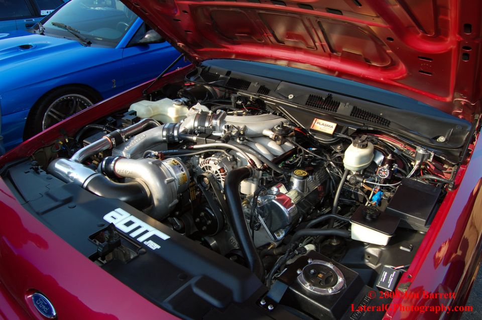 Engine bay of a red car with a turbocharger and a blue car in the background