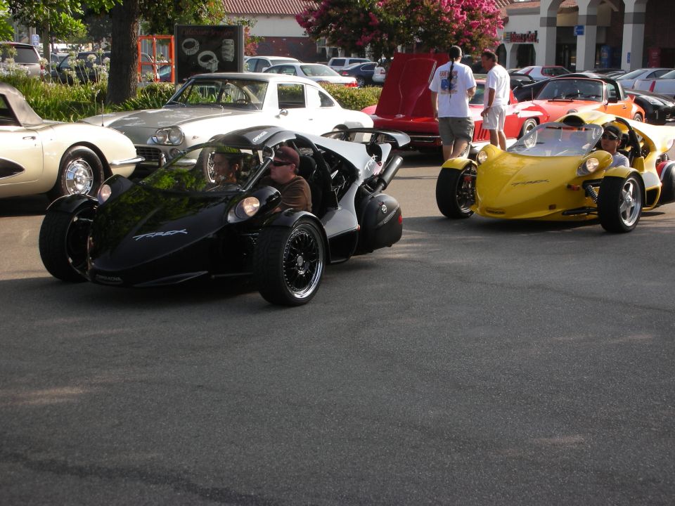 Black and yellow three-wheeled T-Rex vehicles being driven at a car show