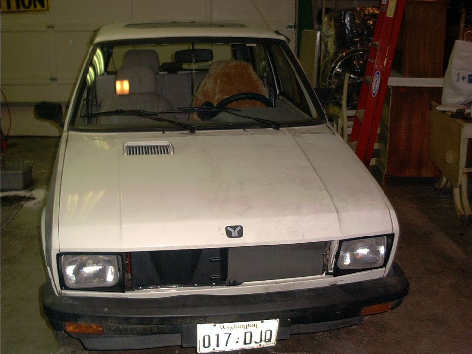 White Yugo hatchback with missing grille, front view in a garage, Washington plate 017-DJQ