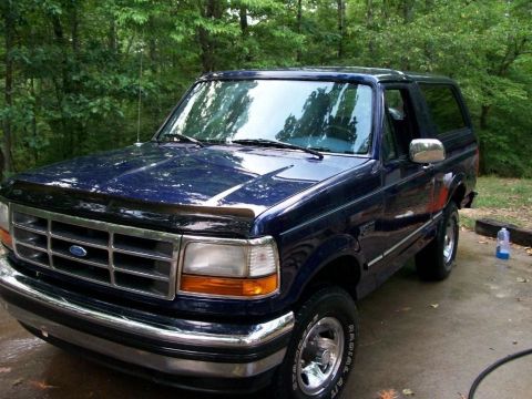 1994 Ford Bronco - A dark blue Ford Bronco is parked on a concrete driveway with trees in the background.