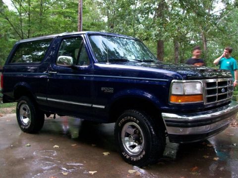 1994 Ford Bronco - Dark blue 1994 Ford Bronco parked on wet pavement with two people visible in the background.