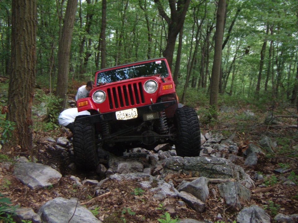 Red Jeep Wrangler navigating a rocky trail in a wooded area. Someone is visible near the front of the vehicle