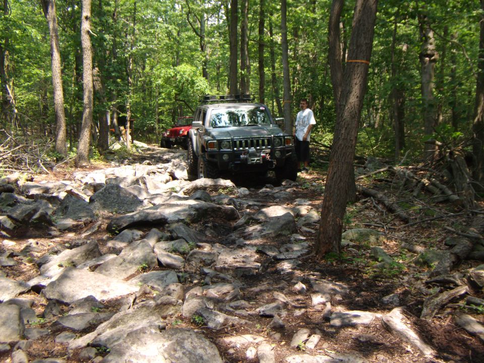 Gray Hummer H3 on rocky forest trail with a person standing beside it