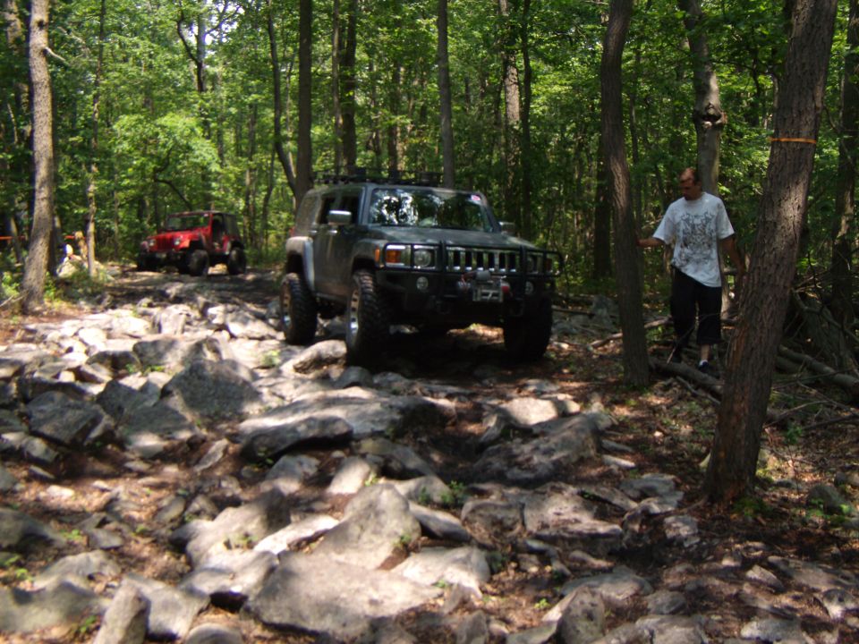 Dark SUV navigating a rocky forest trail with a red Jeep in the background