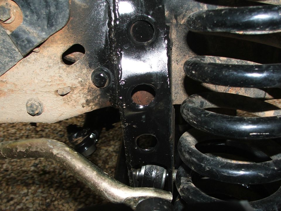 Close-up of a black metal bracket with holes, a black coil spring, and silver tie rod end