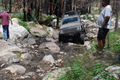 1996 Ford Bronco - A dark-colored Jeep Cherokee XJ is navigating a rocky, muddy stream bed with a person standing on the right watching. Another person is wal…