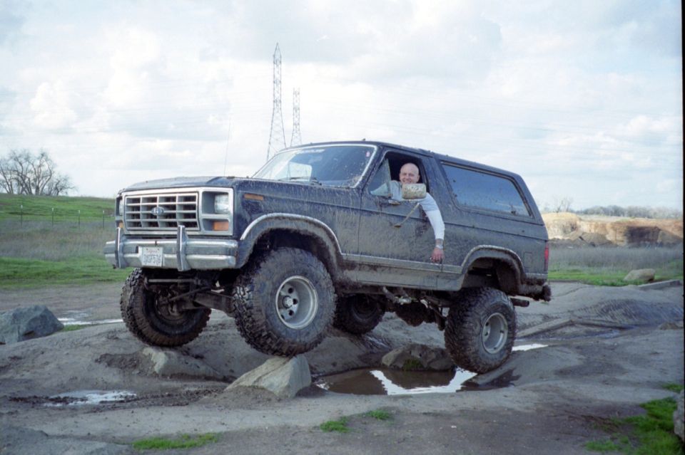 Lifted blue Ford Bronco on large mud tires crossing a rocky puddle with a driver waving