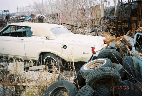 1969 Lincoln III - A white convertible with a tan top sits in a junkyard, surrounded by debris and other vehicles.