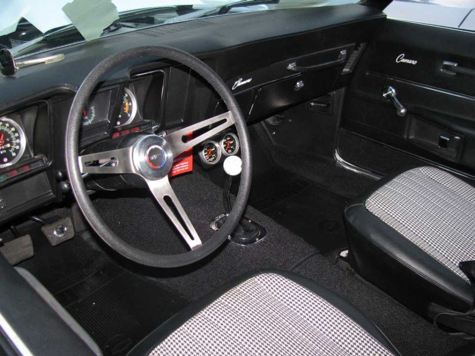 Interior of a classic car with a steering wheel, gauges, and houndstooth seats. "Camaro" text visible.
