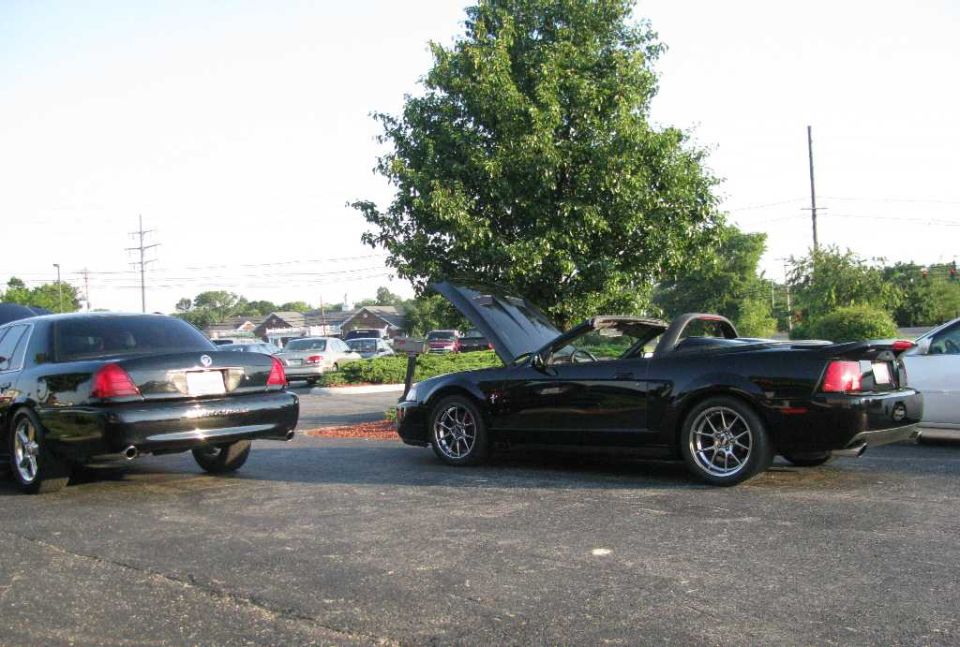 Black convertible Mustang with hood open parked next to a black sedan.