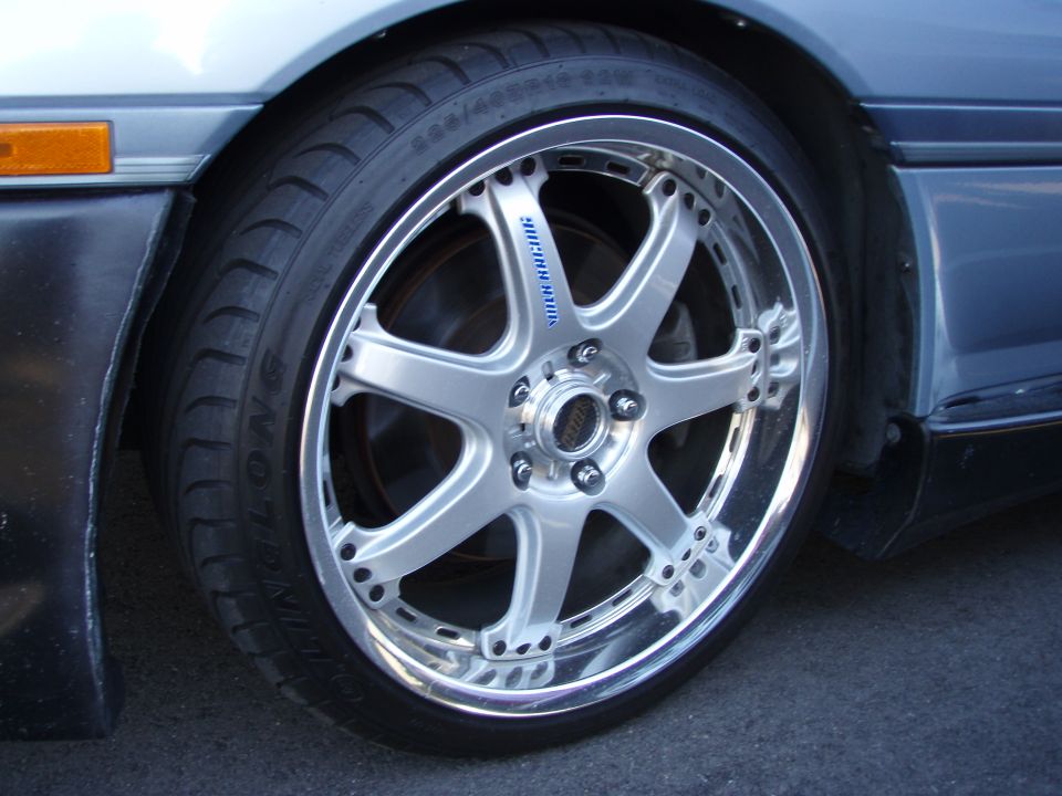 Close-up of a silver multi-spoke aftermarket wheel and tire on a light blue car.