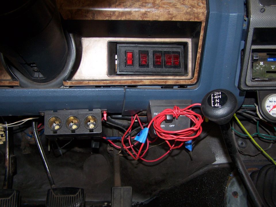 Interior of a vintage Ford Bronco dashboard with toggle switches, gear shifter, and cigarette lighter adapter.