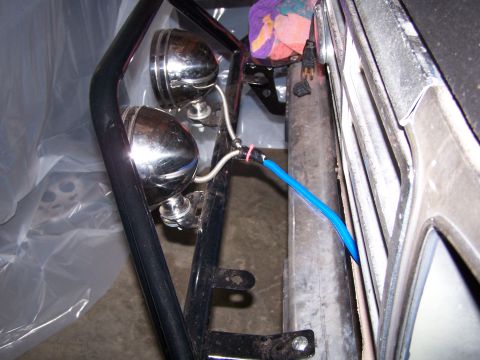 1986 Ford Bronco - Two chrome aftermarket fog lights are mounted to a black roll cage in front of a vehicle. A blue wire loom is routed near the vehicle's fra…