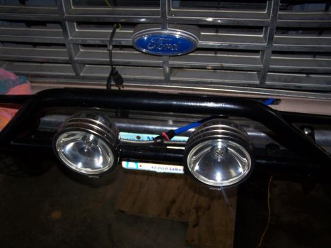 1986 Ford Bronco - A close-up view of the front grille and bumper of a vehicle, featuring two aftermarket fog lights mounted on a black bull bar. A blue Ford…