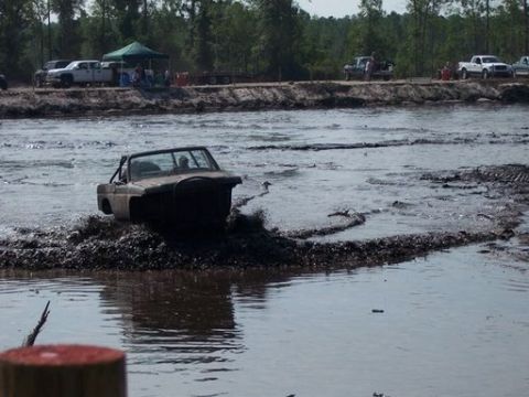 1989 Ford Bronco - A modified dark-colored off-road vehicle is driving through a muddy track, kicking up water and mud. Several trucks and people are visible…