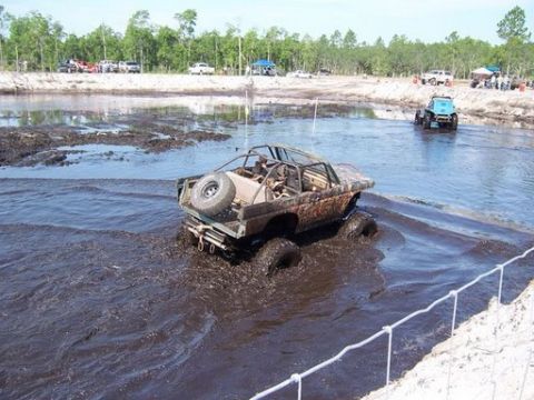 1989 Ford Bronco - A heavily modified off-road Bronco is navigating through a deep mud pit, with another blue off-road vehicle visible in the background. The…
