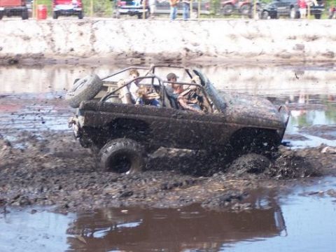 1989 Ford Bronco - A muddy, open-top off-road vehicle is navigating through a deep mud pit, with several people inside. The vehicle is heavily covered in mud,…