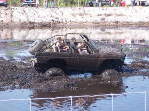 1989 Ford Bronco - A muddy, open-top 1989 Ford Bronco is stuck in a mud pit with several people inside. The vehicle is covered in mud, with its tires submerge…