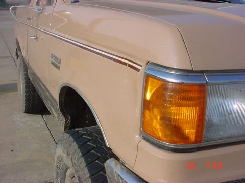1989 Ford Bronco - Close-up of the front fender and headlight assembly of a tan 1989 Ford Bronco, showing its distinctive body style.