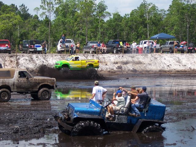 Mud bog event with a mud-covered Ford Bronco, a blue open 4x4 full of riders, and a lime-green lifted truck on the berm