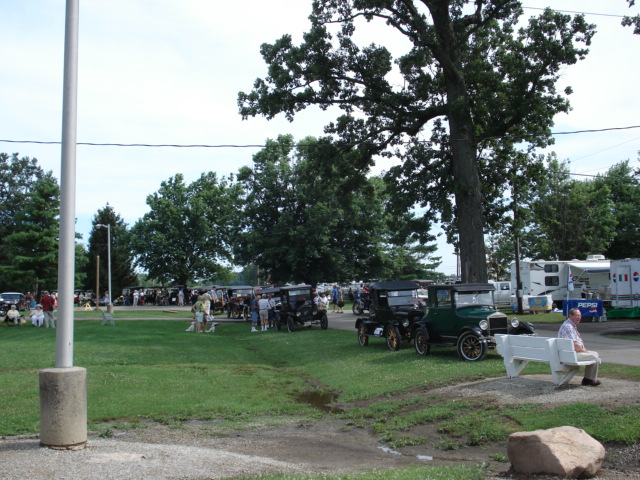 Several dark-colored touring cars from the 1910s parked on grass, one green car in foreground