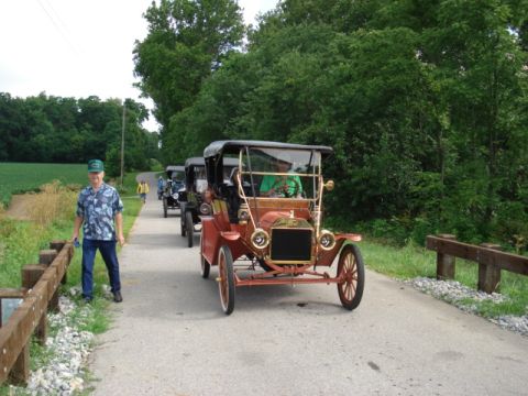 1909 Ford Model T - A line of vintage Ford Model Ts, including a red one in the foreground, travel down a country road.