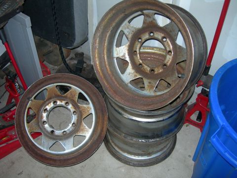 1985 Ford Bronco - Several old, rusty wheels are stacked in a garage, with a blue bucket and red jack stands visible.