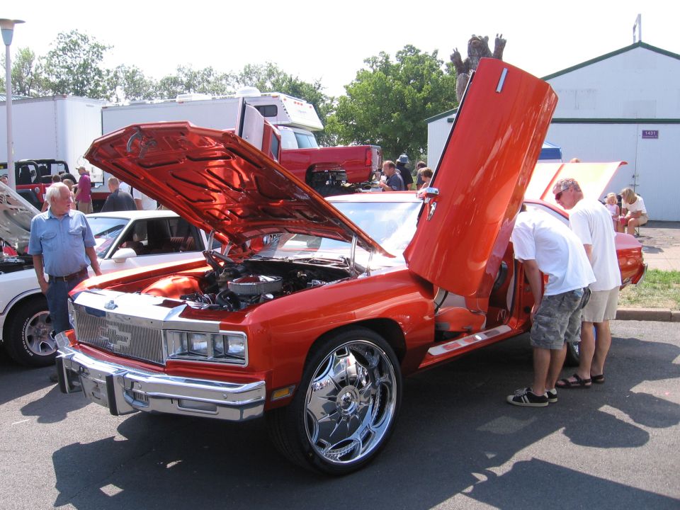 Orange custom Chevrolet with vertical lambo door open and chrome wheels at a car show