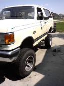 1987 Ford Bronco - This lifted 1987 Ford Bronco is shown from a three-quarter front perspective, highlighting its substantial tires and elevated stance.