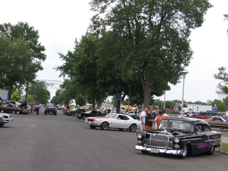 Black 1955 Chevrolet with exposed engine, parked on street with other classic cars and people