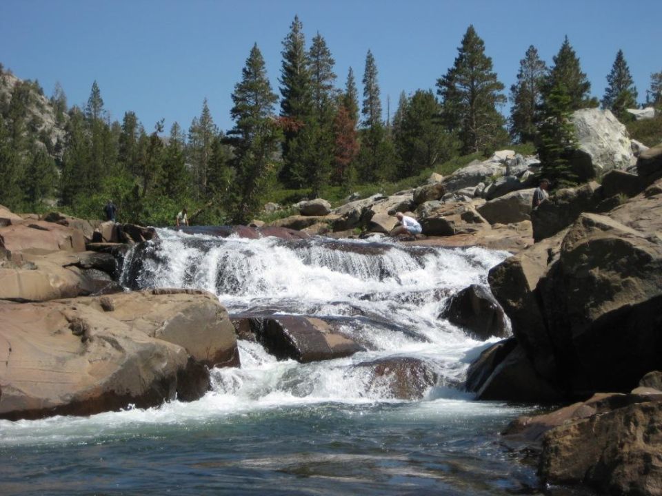 Rocky waterfall with trees and blue sky. Water flows over rocks into a pool. Two people are visible near the top