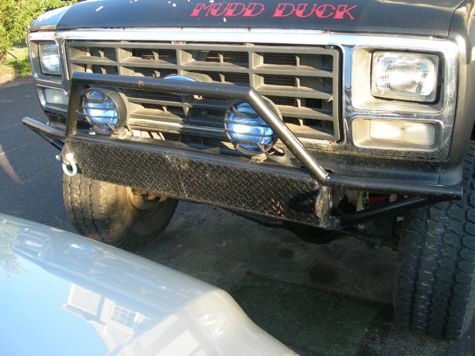 Front end of a black Ford truck with "MUDD DUCK" painted on the hood, custom bumper, and blue auxiliary lights. Grille suggests 1980s model