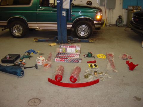1996 Ford Bronco - A collection of new parts, including shocks and springs, are laid out on the floor next to a green Ford Bronco in a garage.