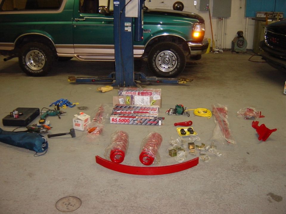 Green 1996 Ford Bronco on lift, with new suspension parts like shocks and springs laid out on the floor