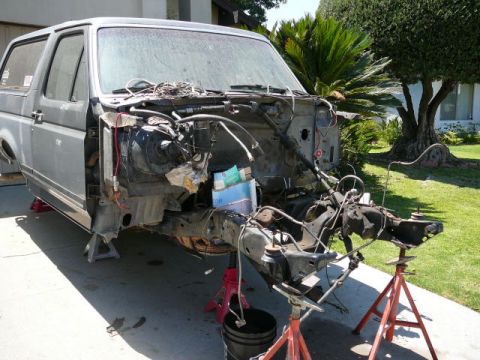 1988 Ford Bronco - Gray Ford Bronco stripped down in a driveway mid-teardown, sitting on red jack stands with the front clip, fenders, grille and bumper remov…