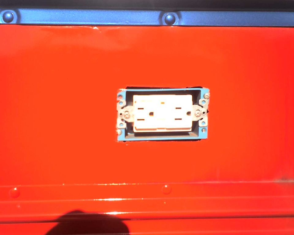 Electrical outlet installed on a red panel with a blue trim piece above. Some rivets are visible