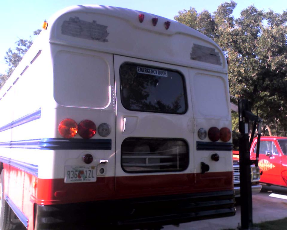 Rear view of a white and red school bus with emergency door visible; a red pickup truck is parked nearby