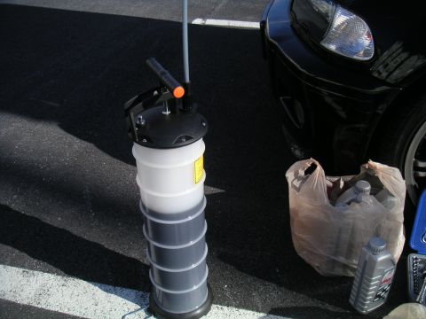 2002 BMW M3 - An oil drain pan sits on asphalt next to a car, with new oil bottles in a plastic bag nearby.