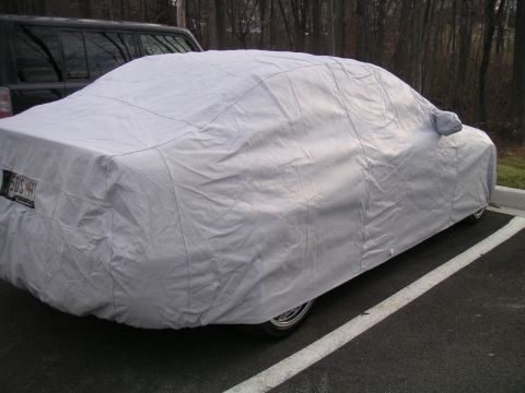 2002 BMW M3 - A car is covered by a grey car cover in a parking lot, with a dark SUV behind it.