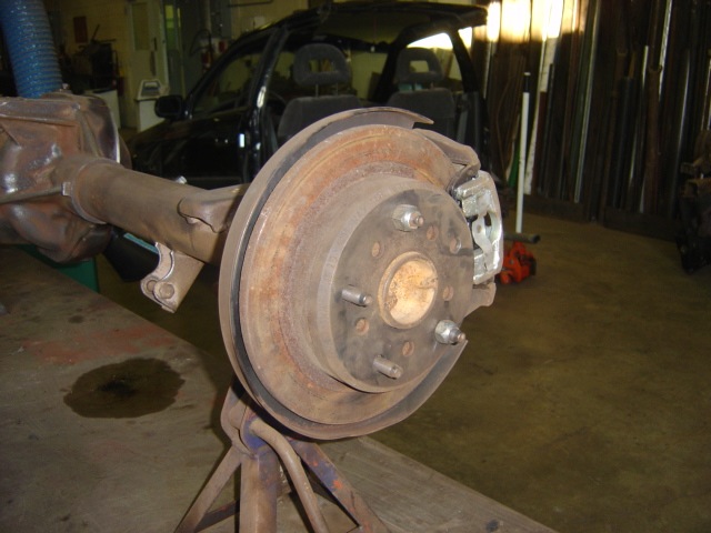 Rear axle assembly with brake rotor and caliper on a stand. A black car is visible in the background