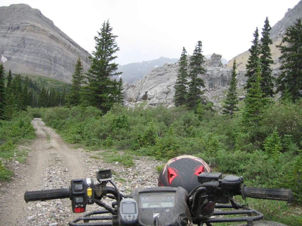 View from an ATV on a rocky dirt road in a mountainous, forested area. A helmet is on the ATV
