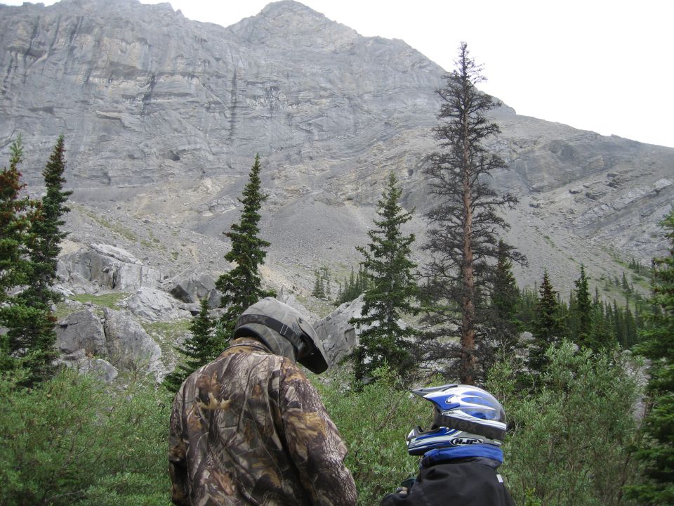 Two people in helmets and camouflage stand in a wooded area with a large mountain in the background